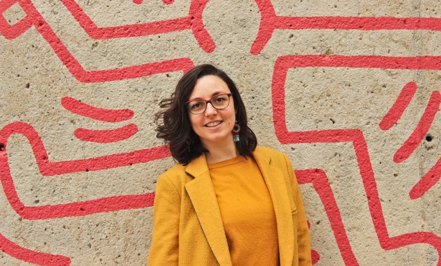 Alice Orrù, freelance copywriter e web writer, indossa un cappotto giallo ocra e sorride di fronte a un'opera di street art di Keith Haring.