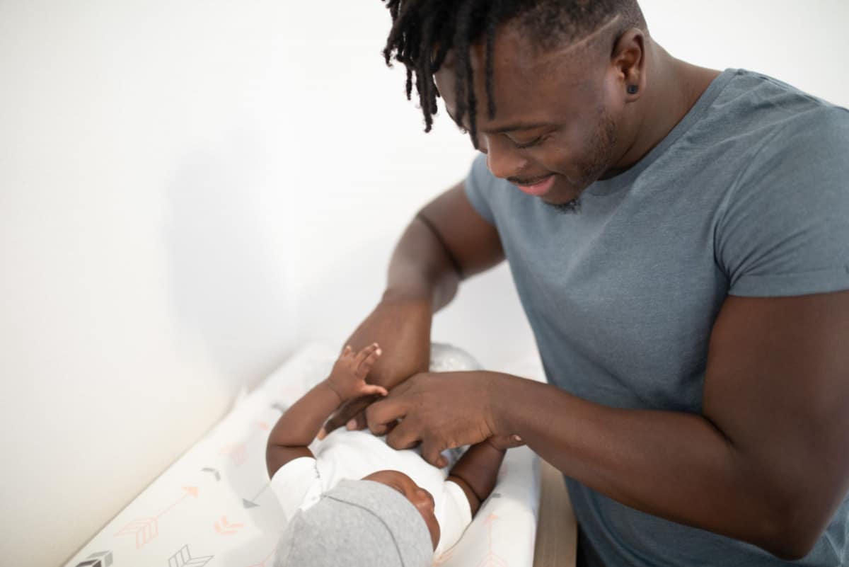 Un uomo afrodiscendente cambia il pannolino a un neonato. Indossa una maglietta grigia mentre il bambino è vestito di bianco.