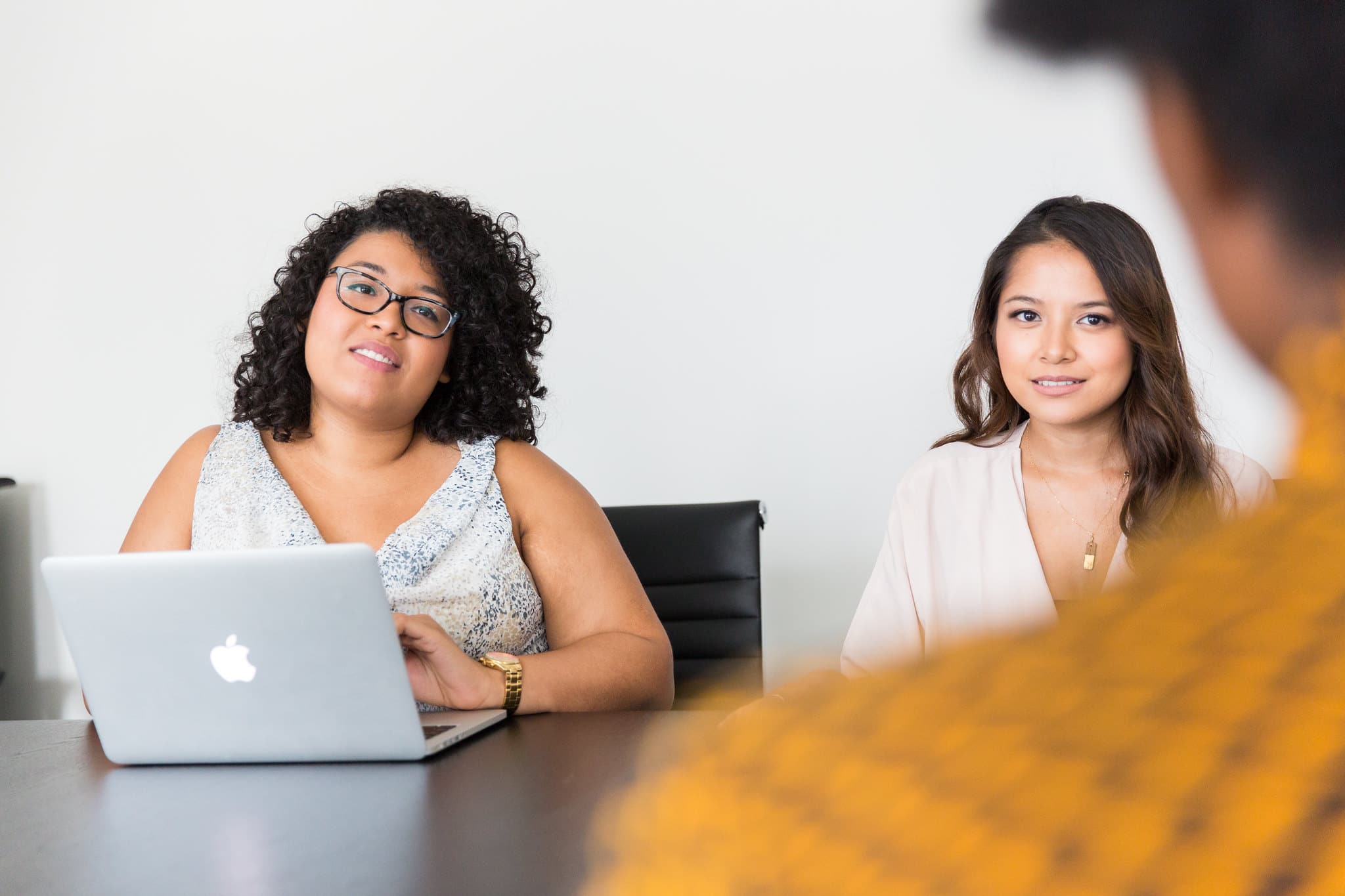 Due donne siedono alla scrivania e sorridono leggermente mentre ascoltano una terza persona che dà le spalle all'obiettivo durante un colloquio di lavoro.