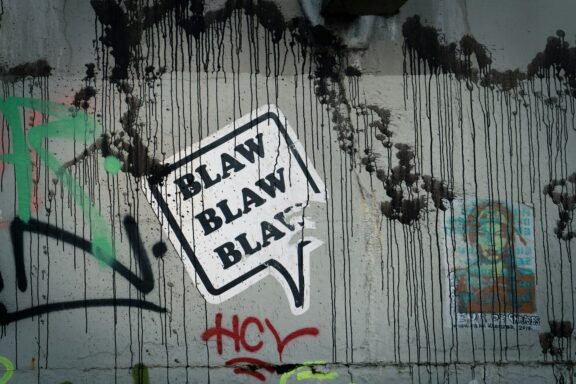 Adesivo bianco a forma di fumetto con la scritta: Blaw Blaw Blaw. È attaccato su un muro pieno di graffiti.
